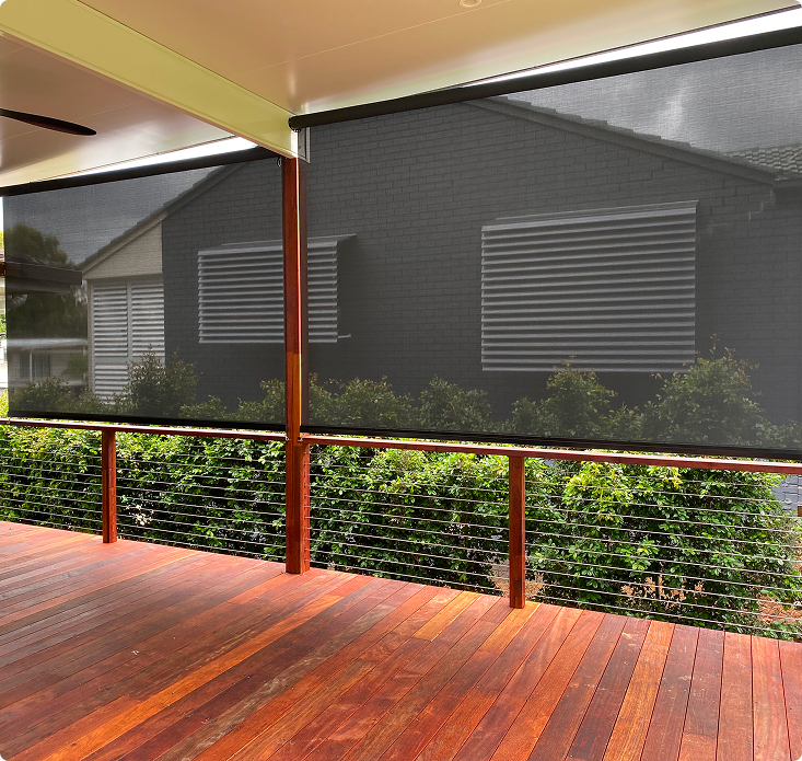 A covered wooden deck with cable railing and black mesh outdoor blinds partly lowered, overlooking green shrubs and a neighboring brick house.
