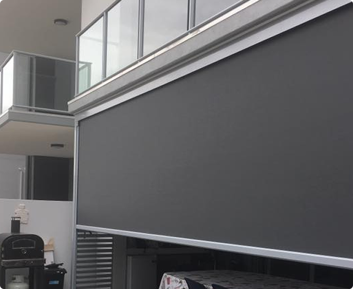 A large dark outdoor roller blind is fully lowered, covering a patio area below a balcony with glass railings. A barbecue grill and laundry rack are visible in the shaded area.