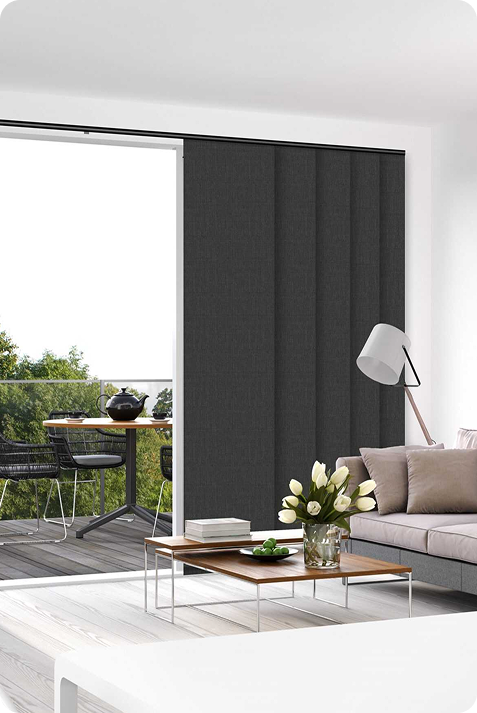 Modern living room with gray sofa, white coffee table, floor lamp, and a sliding glass door with dark vertical blinds, leading to a balcony with outdoor furniture and greenery visible.
