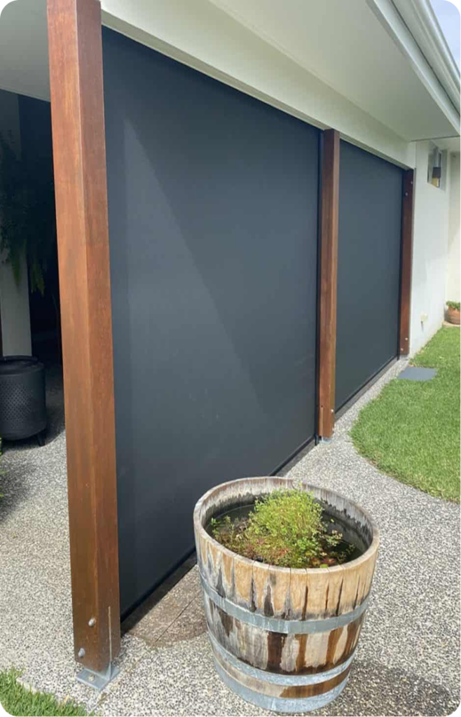 A wooden barrel planter with green plants sits on a patio in front of two large black retractable screens attached to a building.