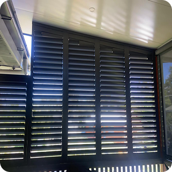 Black louvered shutters with sunlight streaming through the slats, casting shadows on a covered outdoor area.