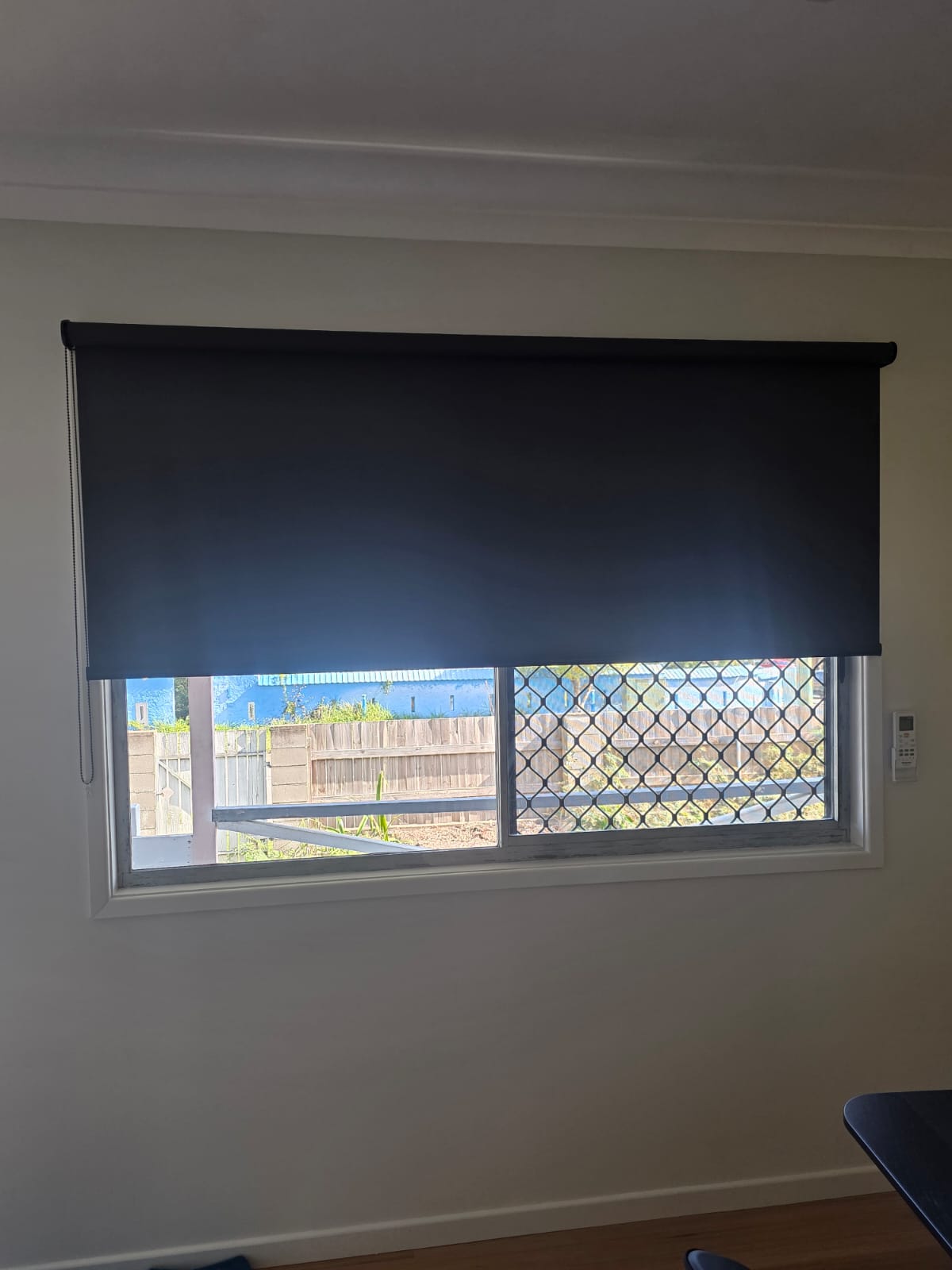 A partially lowered black roller blind covers a window with a security screen, looking out to a backyard and wooden fence.