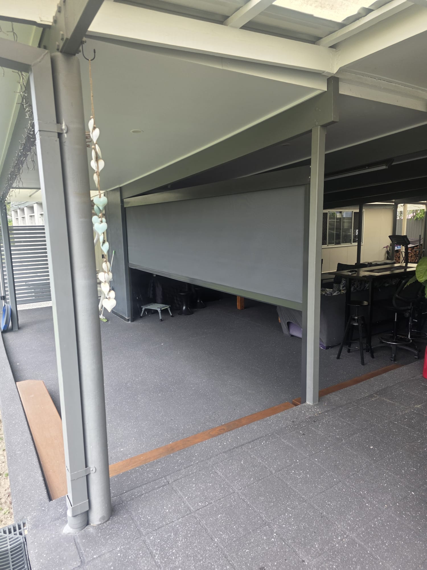 A covered outdoor patio area with a large gray retractable shade partially lowered, supported by metal beams and furnished with tables and chairs.