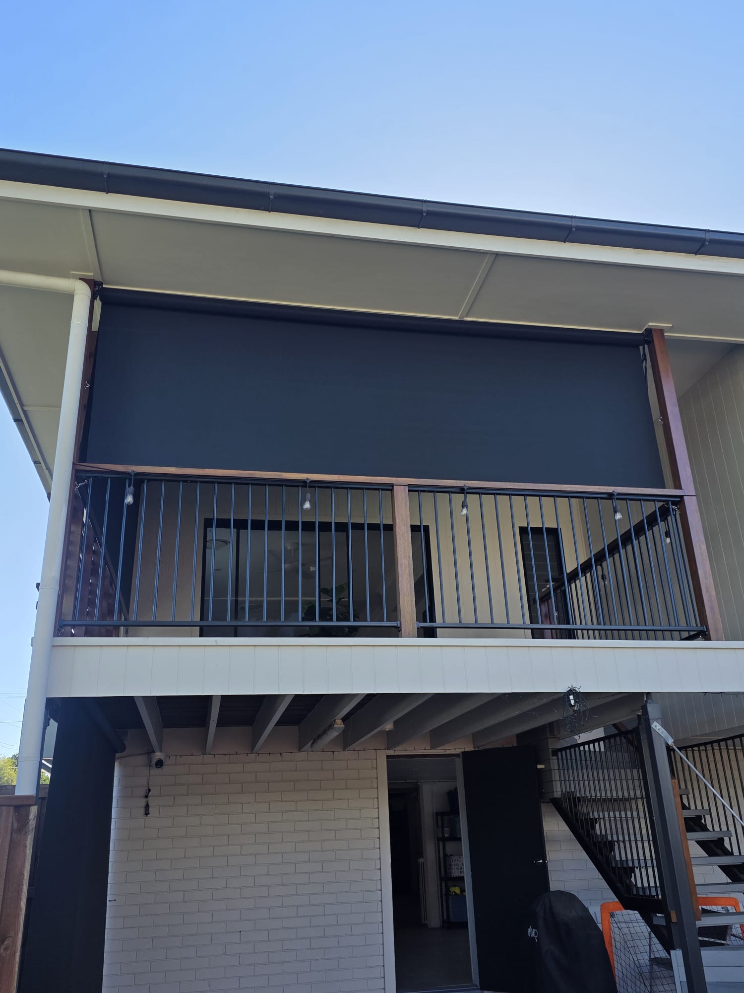 A two-story house with a covered balcony featuring a black roller shade, metal railing, and an external staircase leading to the lower level.