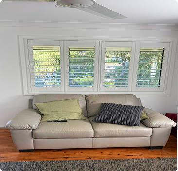 Beige leather sofa with three cushions and a remote on it, positioned in front of four white window shutters in a bright room with wooden flooring.