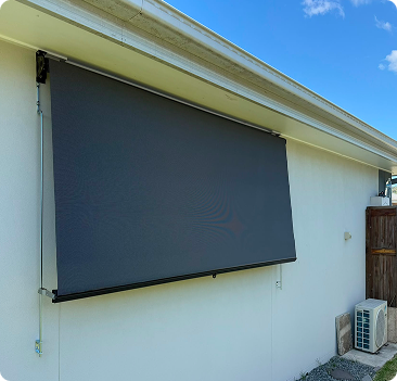A dark outdoor roller shade is mounted on the exterior wall of a light-colored building under a clear blue sky.