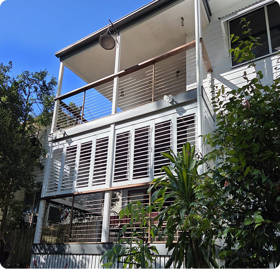 Two-story house with a covered balcony, cable railing, and white louvered shutters on the lower level, surrounded by trees and plants under a clear blue sky.
