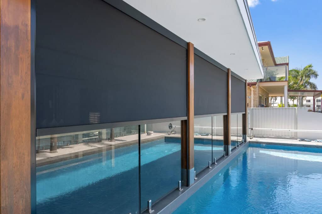 Outdoor roller blinds partially cover glass panels next to a modern swimming pool under a covered patio, creating a stylish and comfortable retreat in Holland Park on a sunny day.