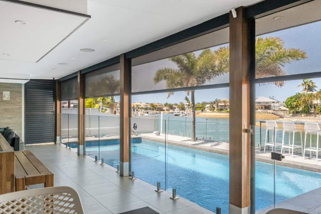 A covered patio with glass fencing overlooks a swimming pool and waterway in Carseldine, with palm trees and houses visible in the background.