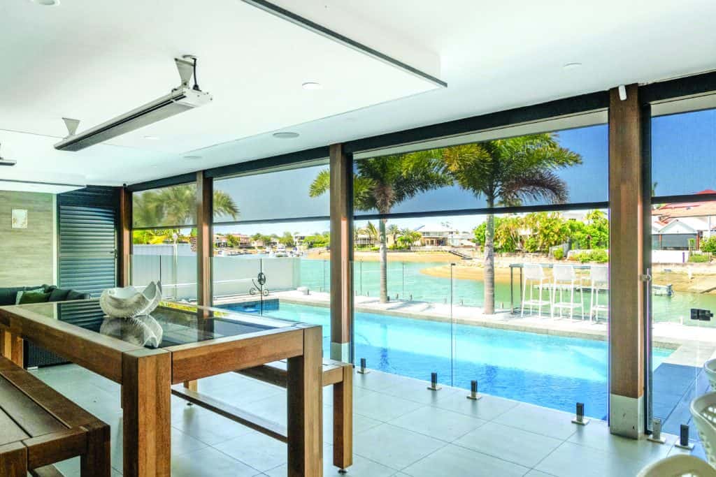Modern indoor-outdoor space in Marsden with glass doors, wooden dining table, pool, palm trees, and a waterfront view under a clear sky.