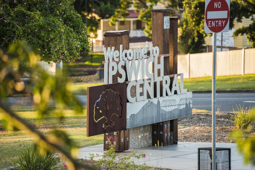 Sign reads “Welcome to Ipswich Central” with a tree graphic, standing near a road with a “No Entry” sign and greenery in the background.