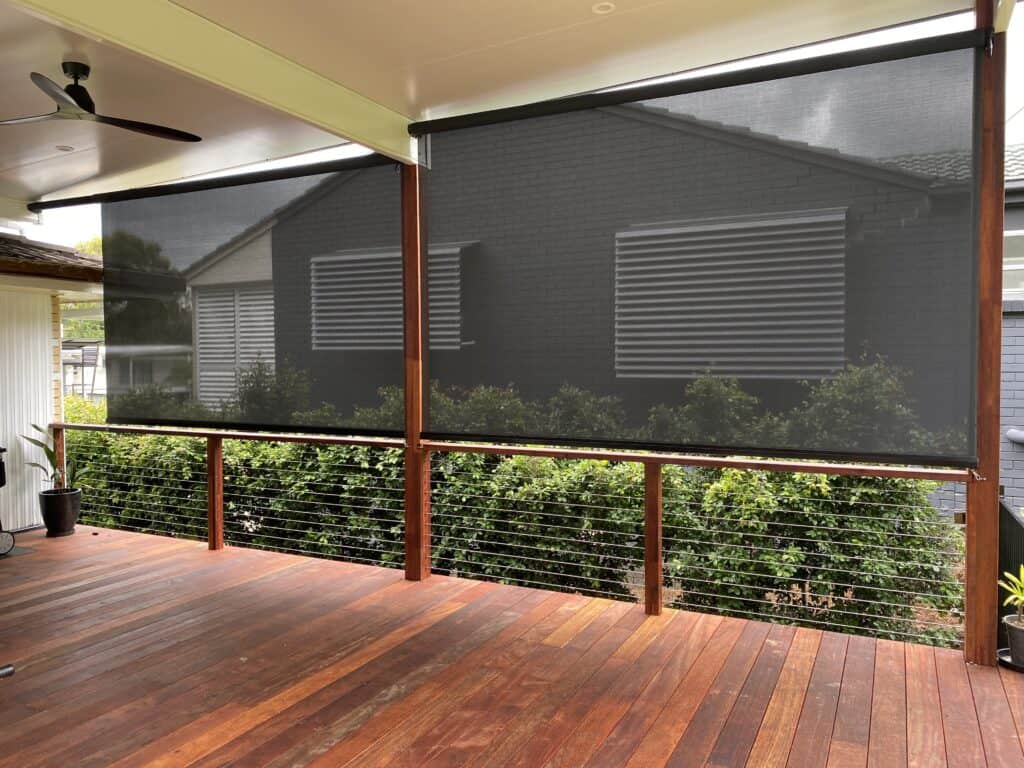 A covered patio in Manly with wooden flooring and black mesh roller blinds partially lowered, overlooking bushes and a neighboring house with grey brick walls and metal window shutters.