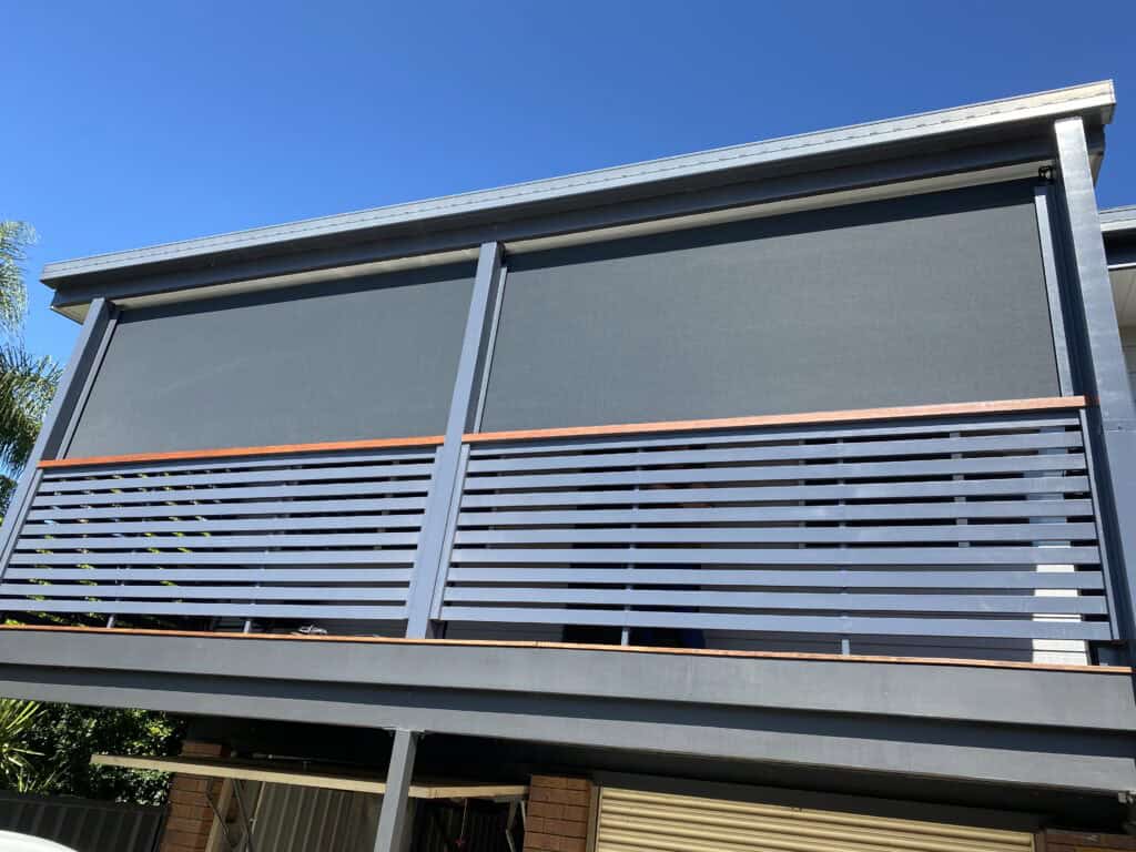 A modern Morayfield balcony with two large outdoor roller blinds fully lowered, horizontal metal railing, and a clear blue sky in the background.