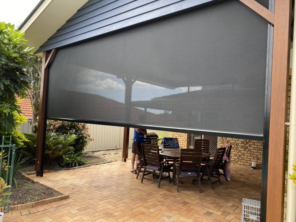 A covered outdoor patio with a wooden dining table and chairs, partially enclosed by a large black mesh outdoor blind.