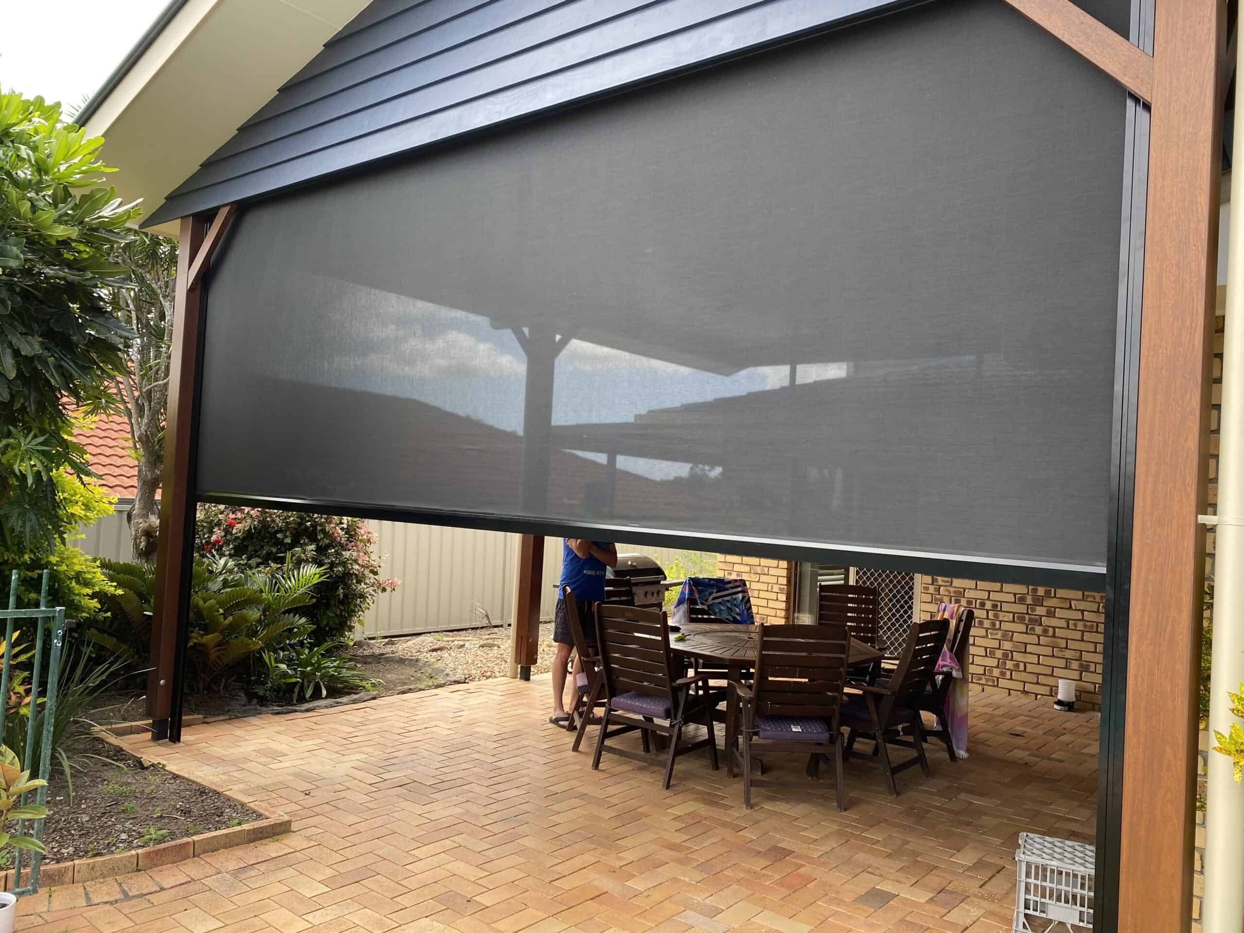A covered outdoor patio area in Beachmere features a wooden dining table and chairs, partially enclosed by a large black mesh screen. A person stands nearby, arranging items on the table.