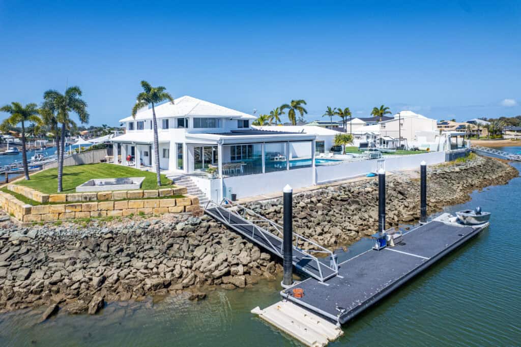 Modern Redcliffe waterfront house with a white exterior, pool, and palm trees, featuring a private dock on a rocky shoreline under a clear blue sky.