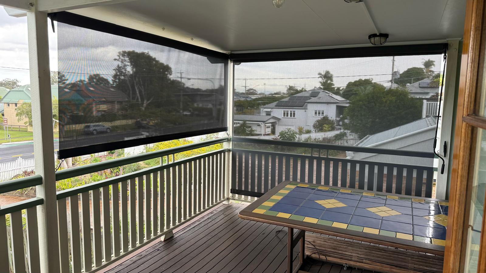 Covered balcony with wooden floor, outdoor table with tile pattern, and mesh screens on two sides; residential neighborhood visible in the background.