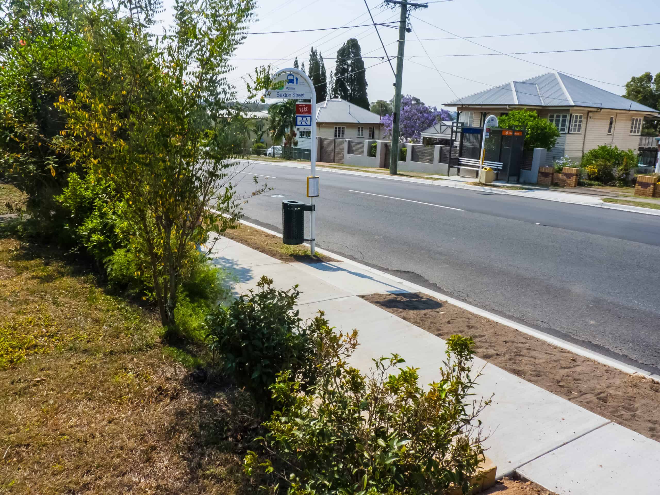 A quiet Tarragindi street with houses, a bus stop sign and a footpath bordered by grass and shrubs on a sunny day.