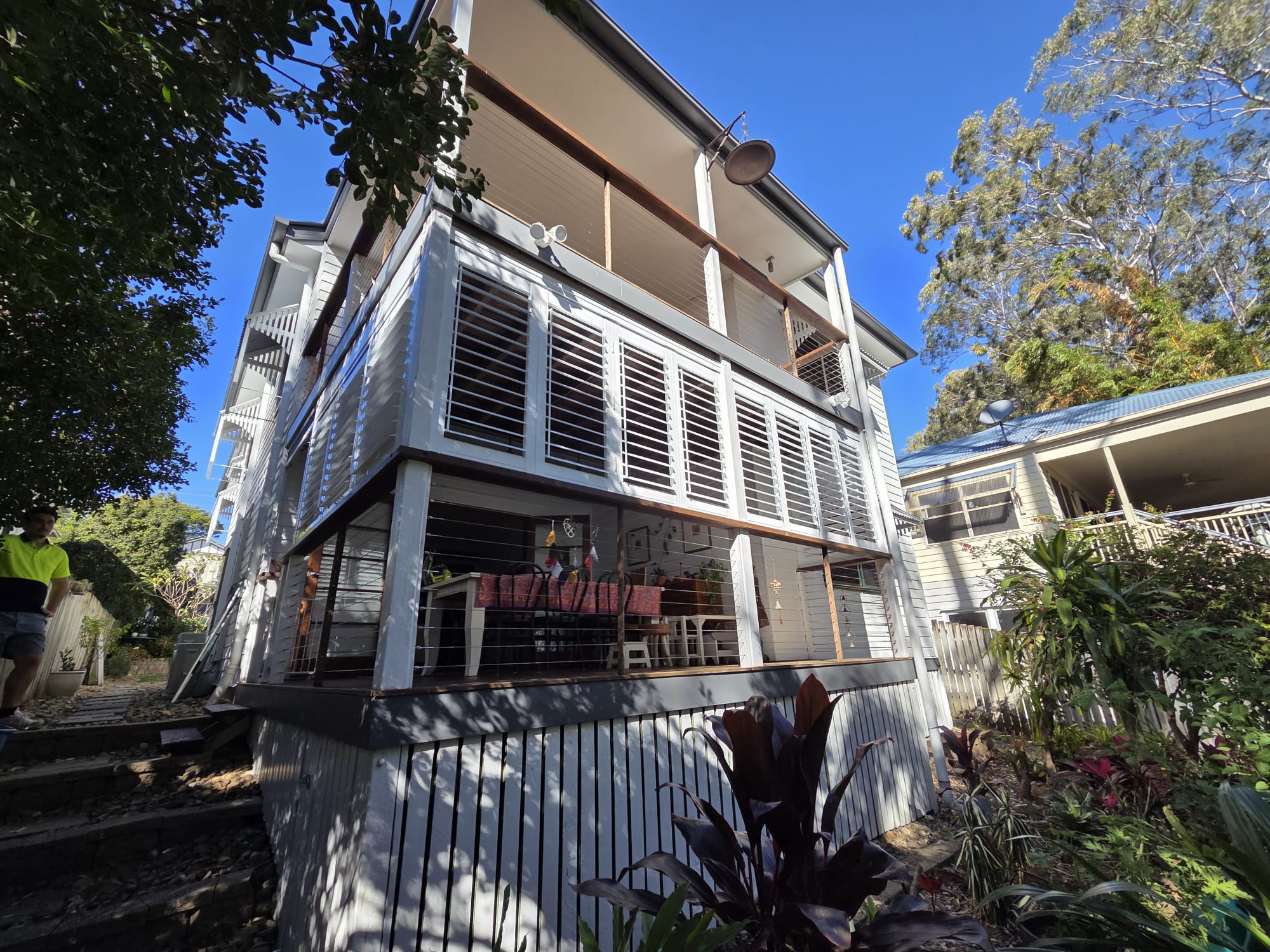 Three-story house with white slatted exterior, multiple balconies, outdoor seating area, surrounded by plants and trees, and adjacent to another house.