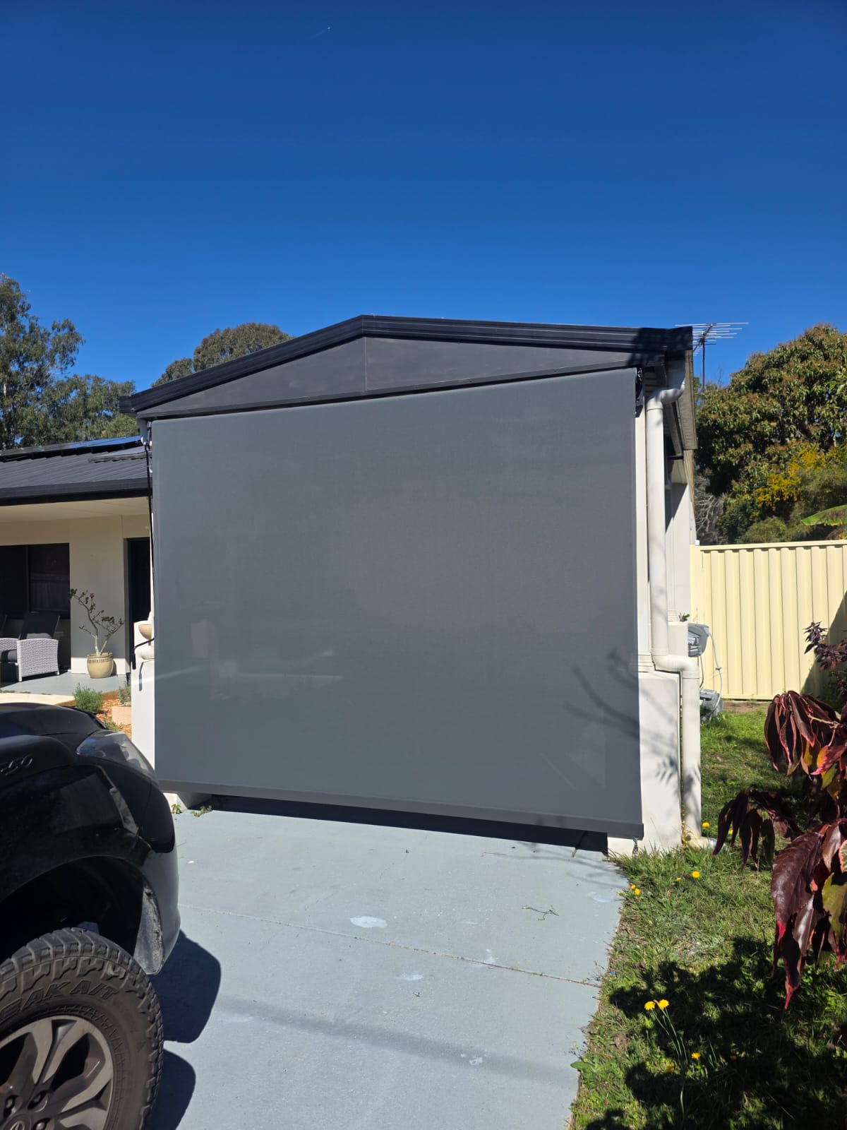 A large, gray outdoor roller blind covers the front of a garage attached to a house, with a black car parked partially in front on a sunny day.