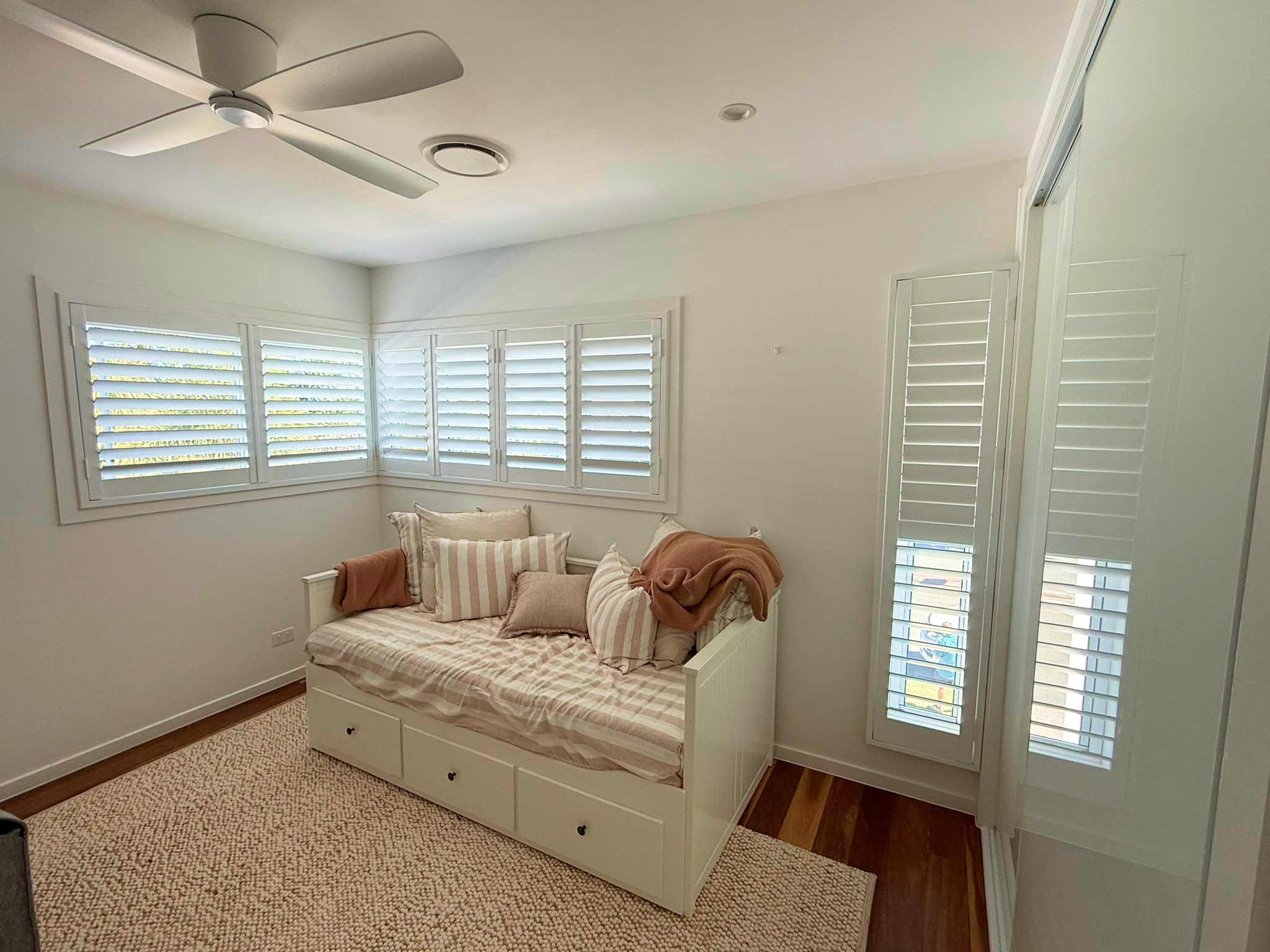 A small bedroom with a white daybed, striped and solid pillows, a beige throw blanket, shuttered windows, a ceiling fan, and a textured rug on wood floors.