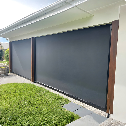 Two large, dark-colored outdoor roller blinds cover the open sides of a patio area attached to a house, adjacent to a grassy lawn and stone walkway.