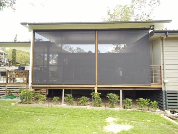 A house with a raised wooden deck enclosed by two large black mesh screens, with small bushes planted along the edge of the deck.