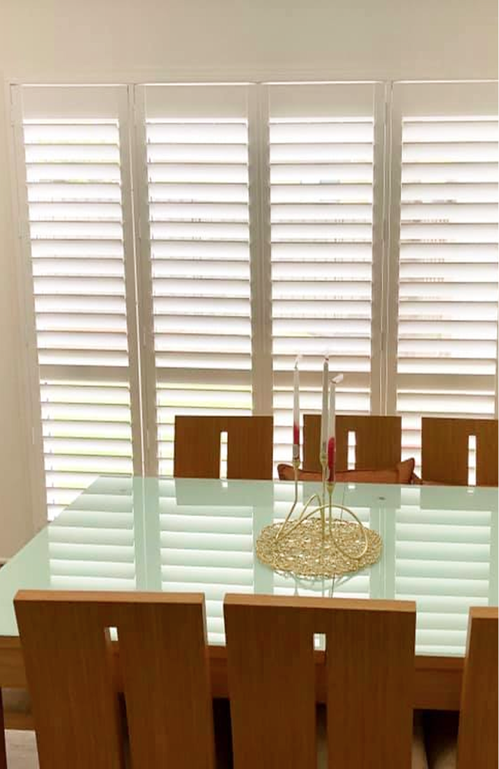 A glass-top dining table with wooden chairs sits in front of large windows with white shutters. Two red candles are on a gold holder in the center of the table.