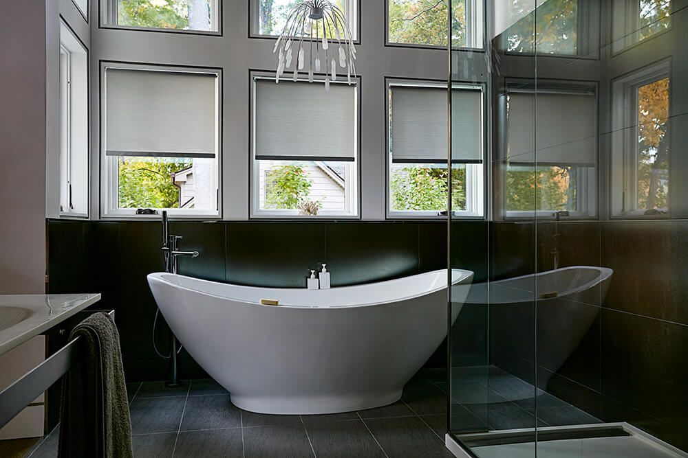 Modern bathroom with a freestanding white bathtub, large windows with blinds, dark tile walls and floors, and a glass-enclosed shower.