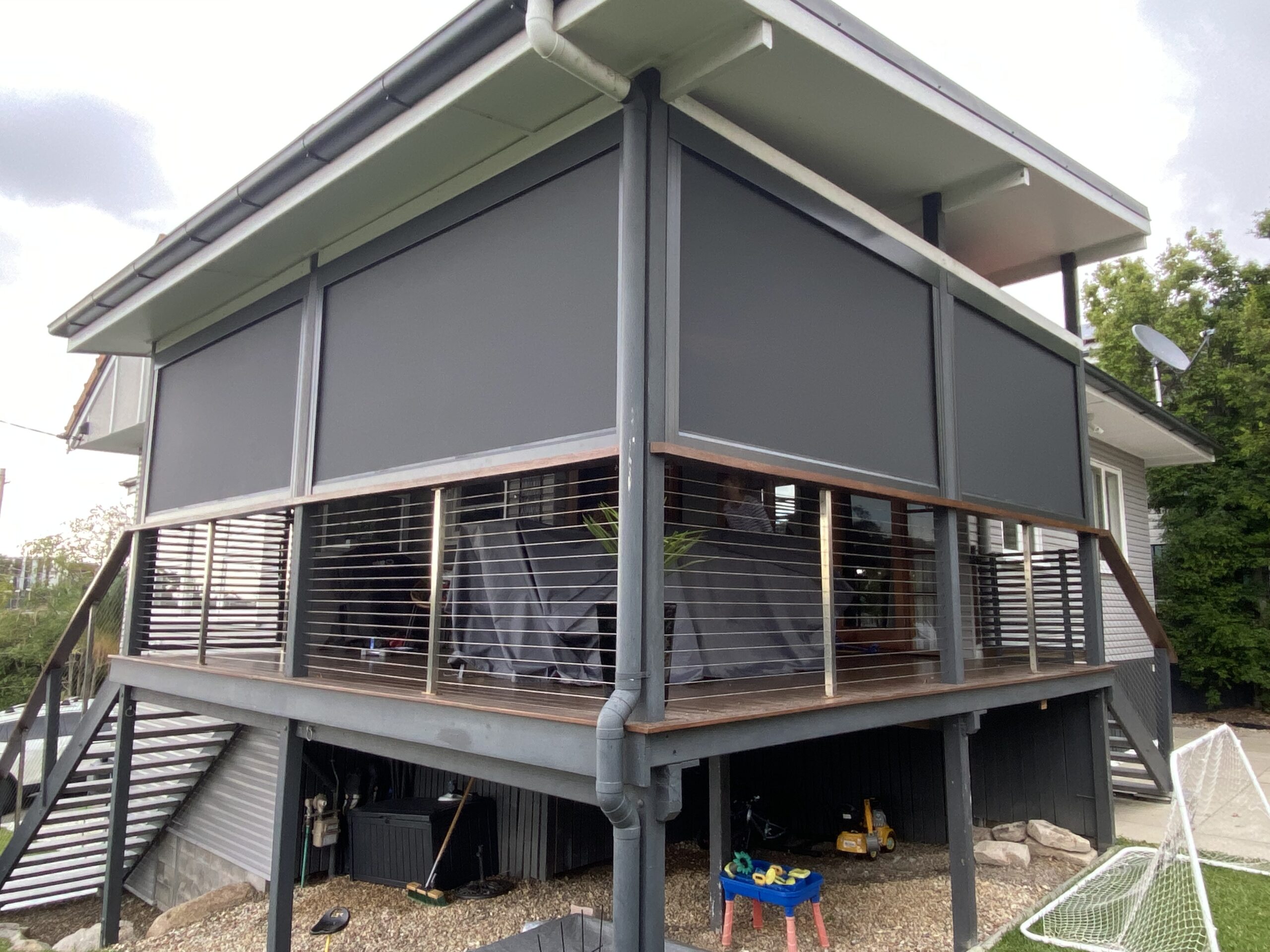 A modern elevated house with an enclosed wraparound balcony featuring dark privacy screens and metal railings. Outdoor items and gravel area are visible underneath the deck.