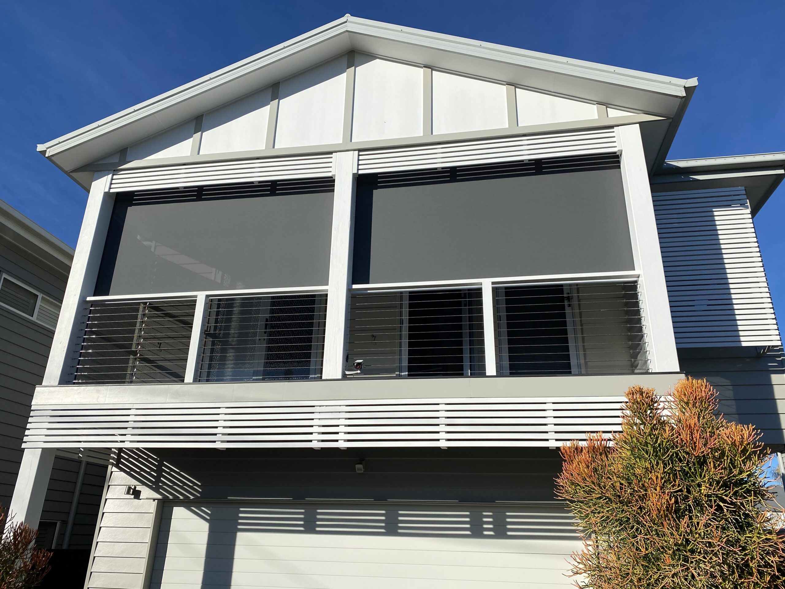 Modern two-story house with a balcony featuring two large, dark roller blinds, metal railings, and a garage underneath. Bushes are visible in the foreground.