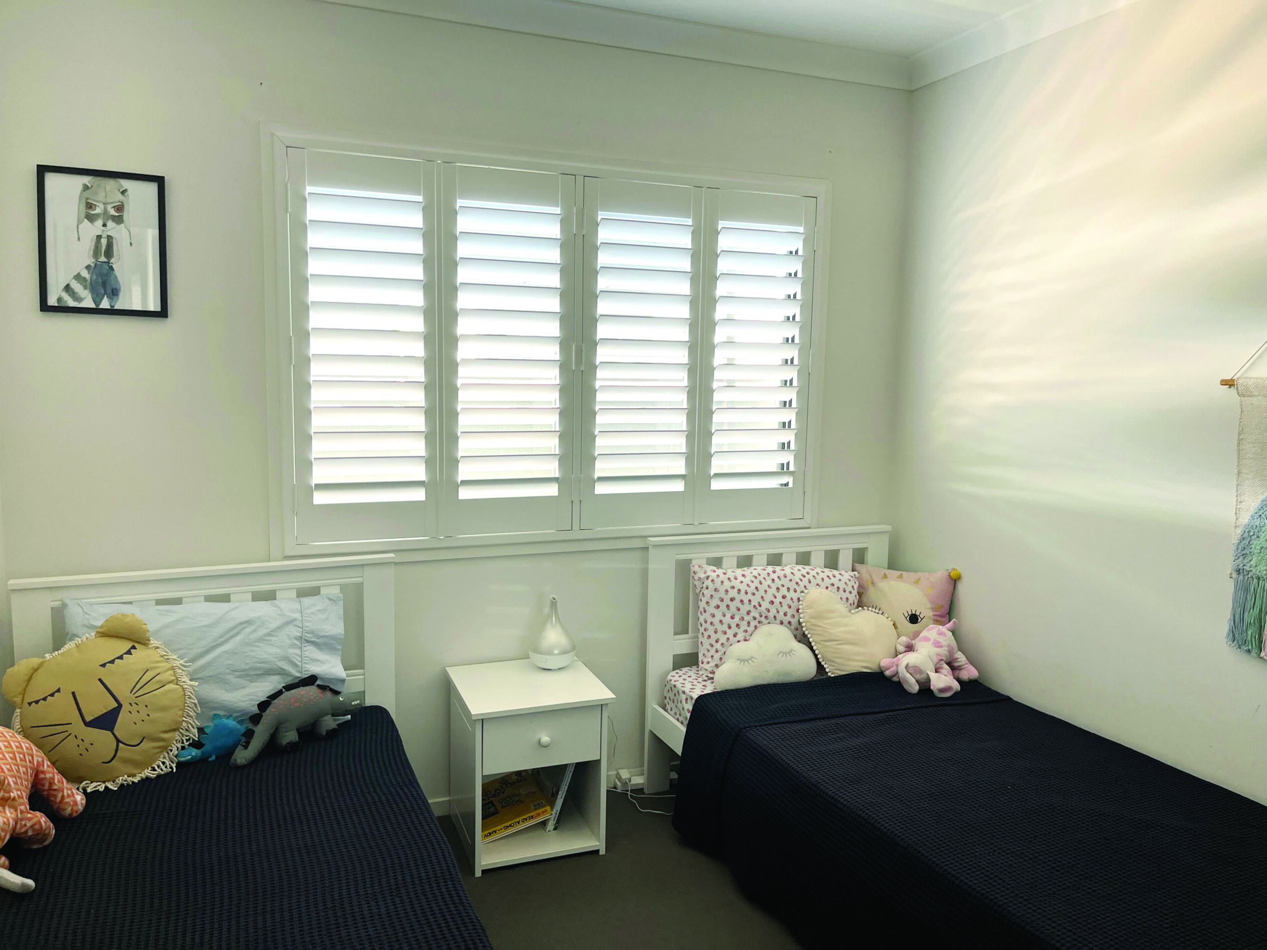 A small bedroom with two single beds, a nightstand, shuttered windows, and stuffed animal pillows on each bed.
