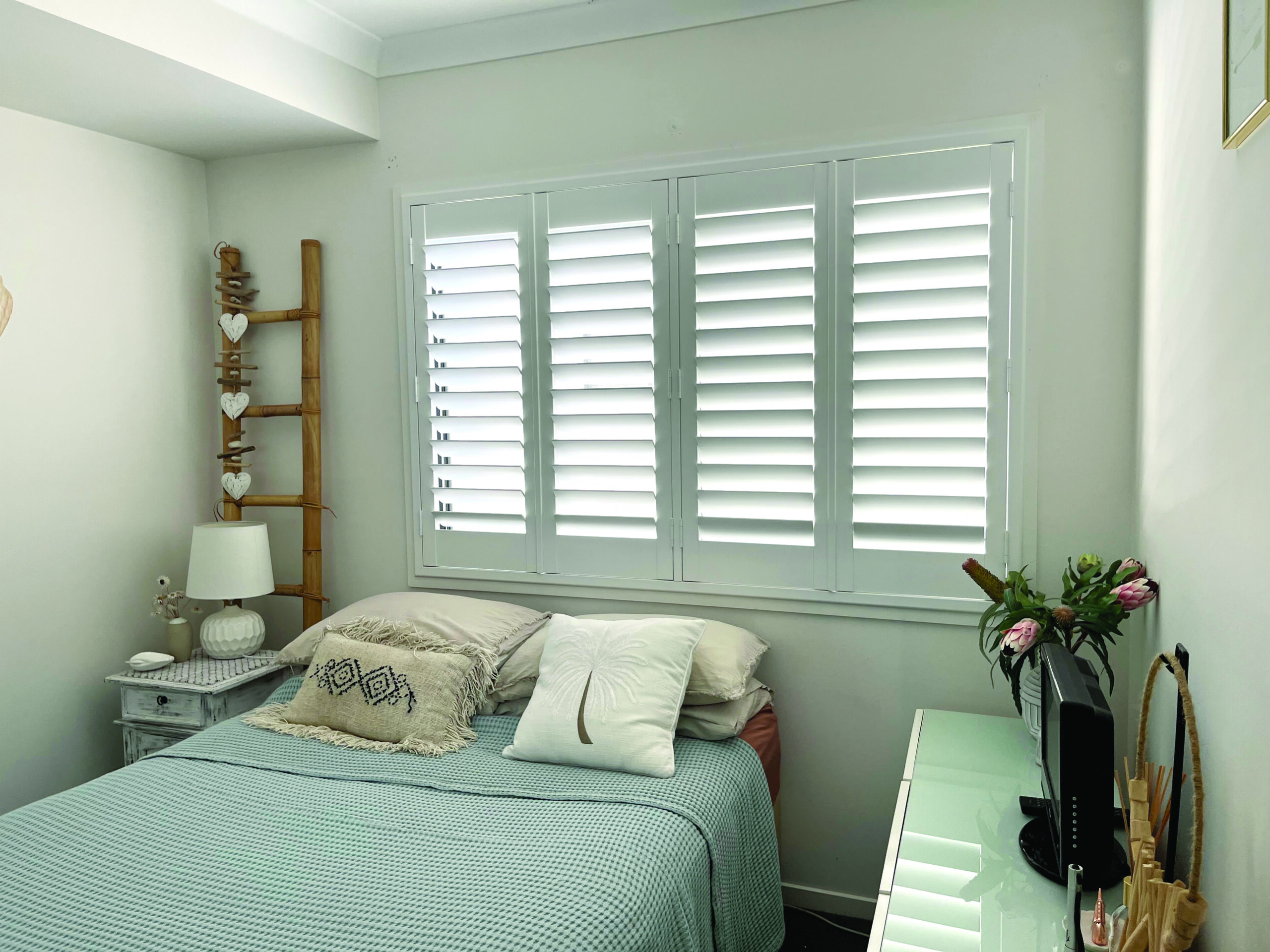 A neatly made bed with pillows and a throw blanket sits by a window with white shutters. A nightstand, lamp, and ladder shelf decorate the left side; a dresser with flowers is on the right.