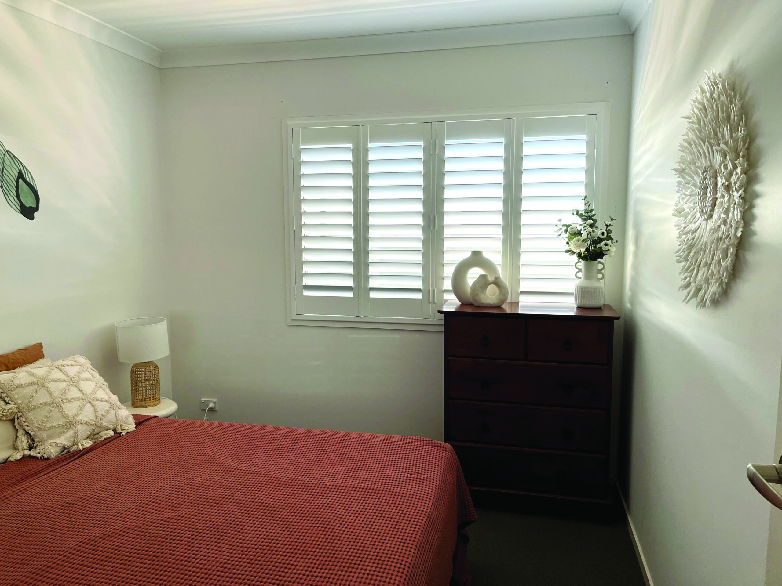 Small bedroom with a bed, red cover, lamp, dark wood dresser, white blinds on window, and decorative items on the wall and dresser.