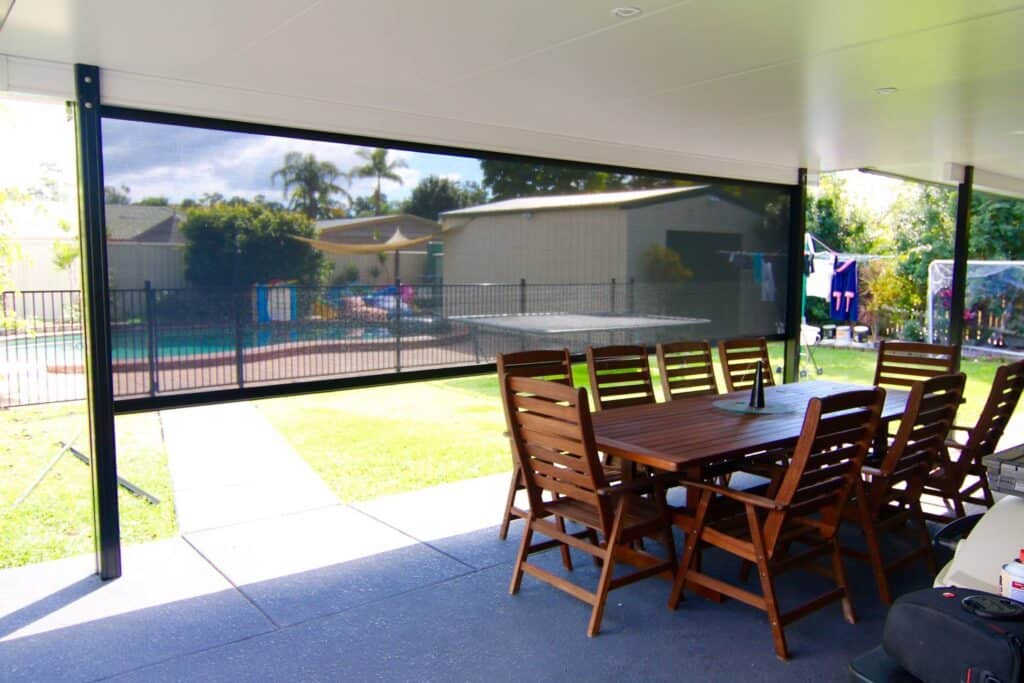 Covered patio area with a long wooden dining table and chairs, overlooking a backyard with a pool, garden, and laundry hanging outside. A shade screen is partially lowered—perfect for relaxed Albany Creek living.