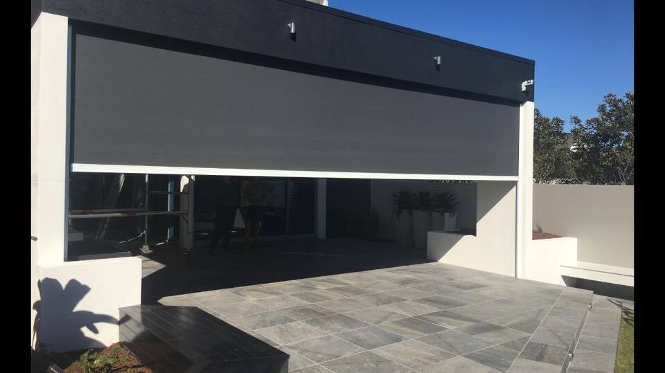 A modern Carseldine house with a large grey retractable patio screen partially lowered over a tiled outdoor area, under a clear blue sky.
