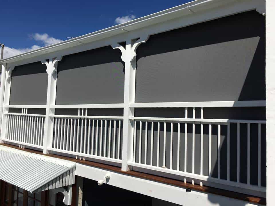 A white Queenslander balcony with vertical railings and decorative supports, fitted with large grey outdoor roller blinds across the upper section.