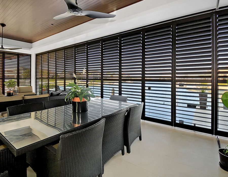 Modern indoor dining area with a glass table, black wicker chairs, and potted plants. Floor-to-ceiling louvered windows provide natural light and a view of the outdoors. Ceiling fans are installed above.