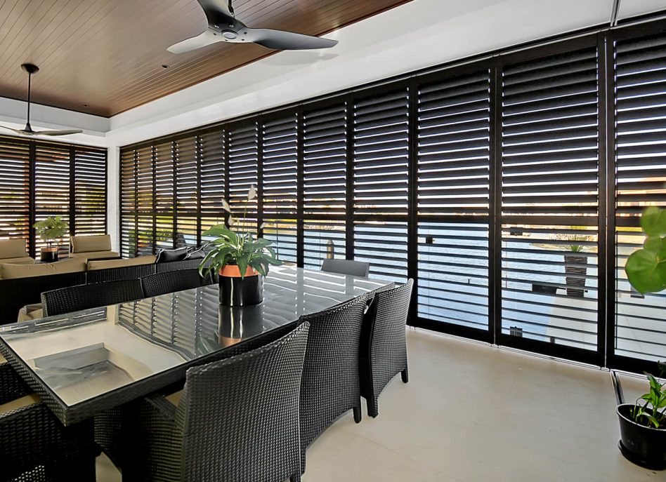 Modern indoor dining area with a glass table, black wicker chairs, and potted plants. Floor-to-ceiling louvered windows provide natural light and a view of the outdoors. Ceiling fans are installed above.