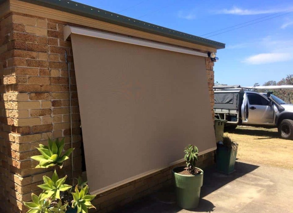 Beige outdoor roller blind on brick wall shading potted plants.