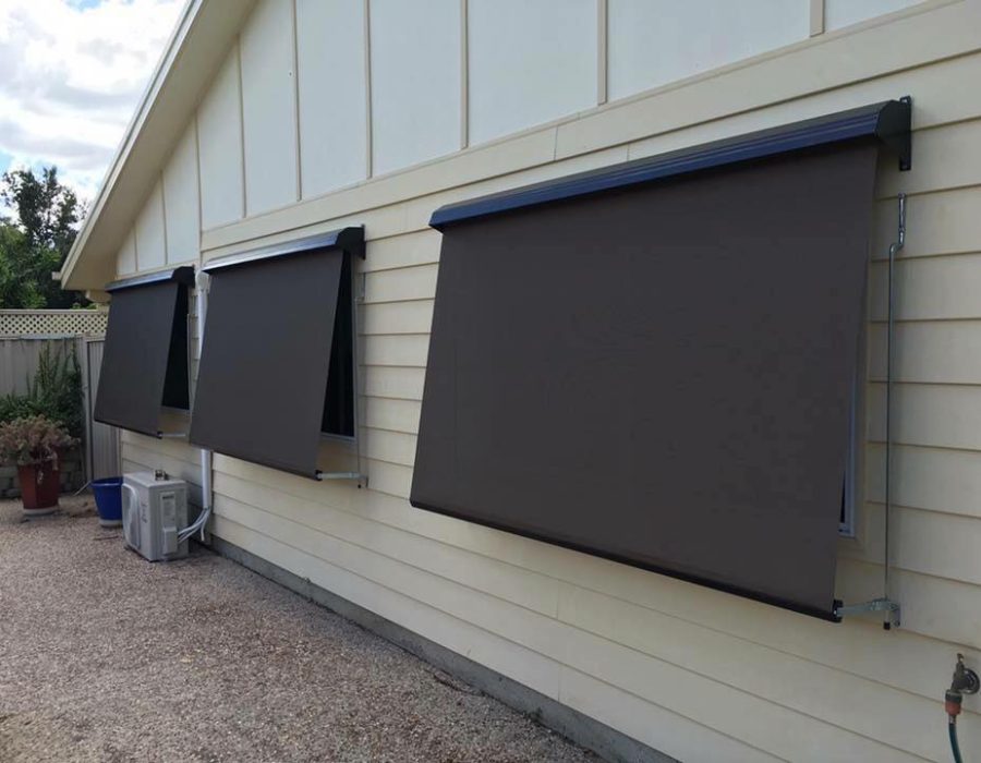 Three black awnings cover windows on the side of a light-colored house. An air conditioning unit is positioned below one of the windows.