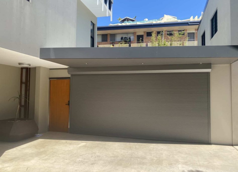 A closed gray roller garage door with a wooden side door stands in a modern Camp Hill residential building courtyard under a clear blue sky.