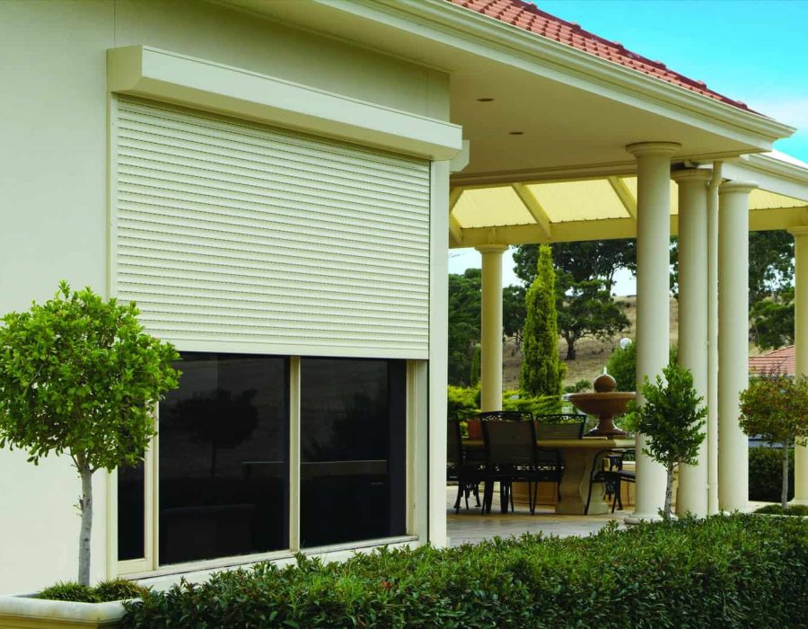 Flagstaff Hill 2 A house exterior with a large window covered by a closed white roller shutter, a hedge, potted plants, and a covered patio area with outdoor furniture.