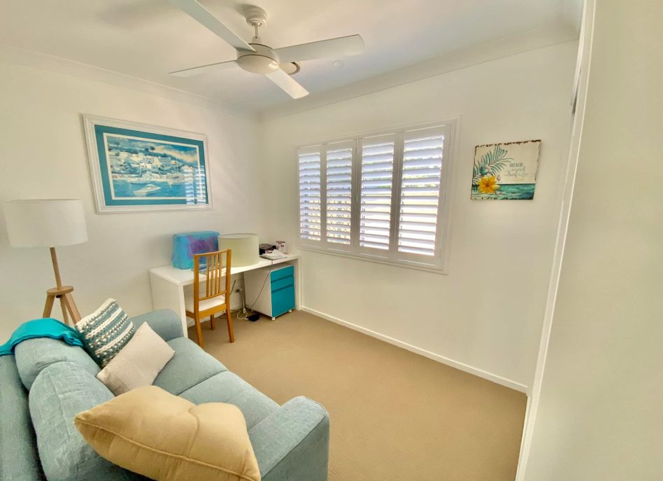 A small home office features a light blue sofa, beige carpet, and white walls. A desk with a chair and lamp sits beneath ocean-themed artwork. Indoor PVC shutters filter sunlight gently while the ceiling fan whirls quietly above.