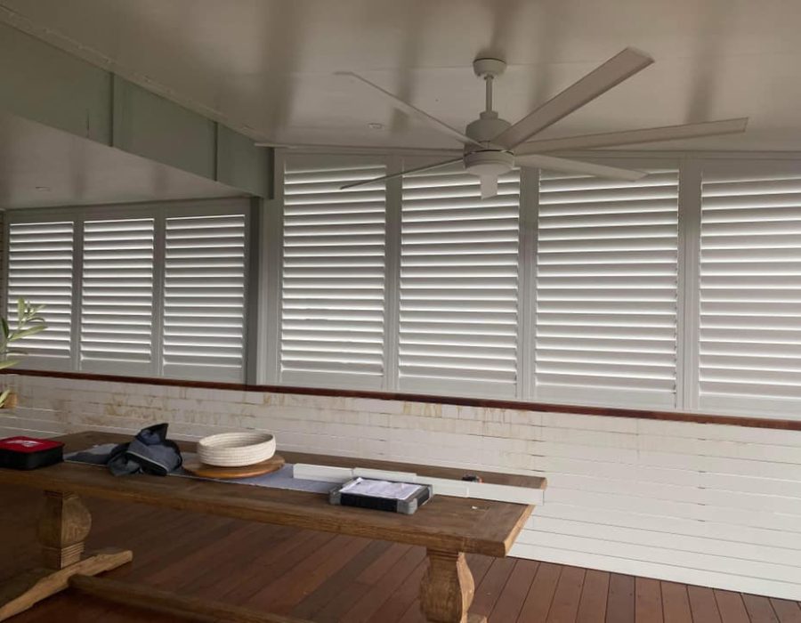 A room with wooden flooring features a long table adorned with a hat, a jacket, and some boxes. White indoor PVC shutters dress the windows, and a ceiling fan is mounted above. The walls are painted white, creating a bright, airy feel.