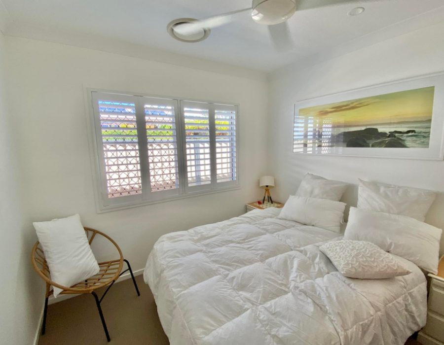 A cozy bedroom with white walls and bedding features a large framed ocean photo above the bed. The window boasts indoor PVC shutters, complementing the plantation style. A wicker chair with a cushion sits in the corner, while a small bedside table holds a lamp.