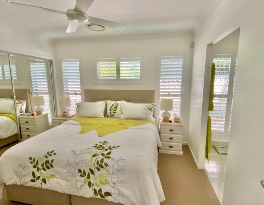 A cozy bedroom with a double bed covered in a white and green floral duvet. The room features mirrored closets, bedside tables with lamps, and a ceiling fan. Indoor PVC shutters adorn the windows, allowing natural light to filter through and create a warm ambiance.