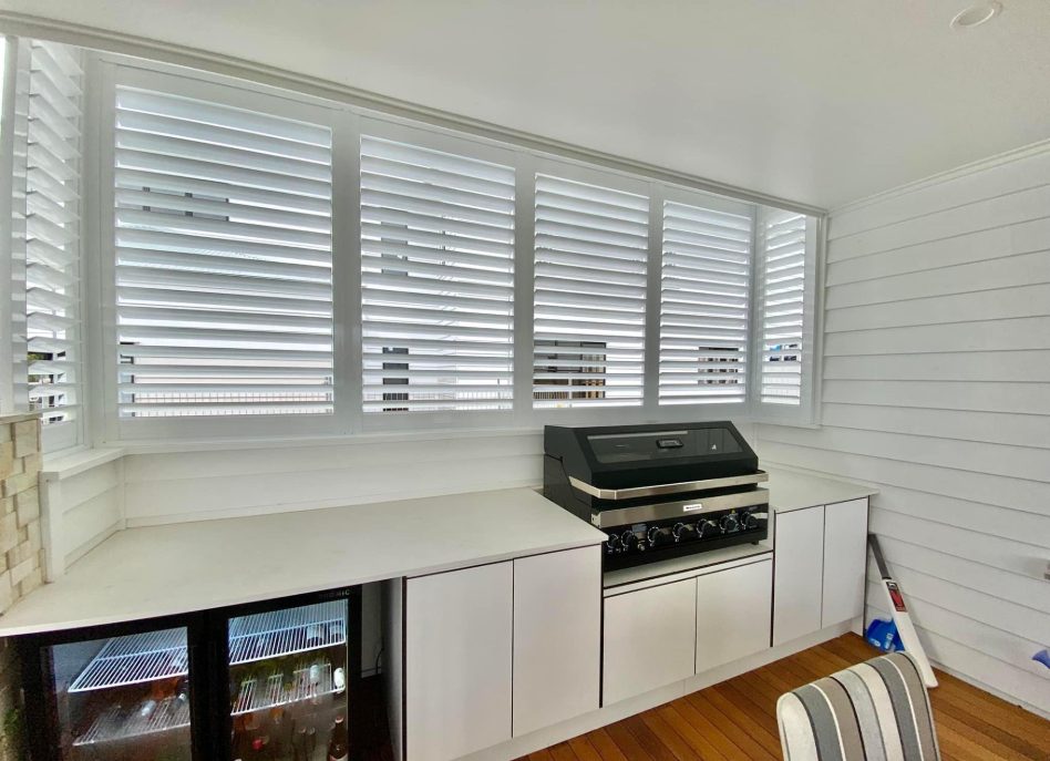 A bright room with white paneled walls and wooden floors features a built-in barbecue grill, a small fridge with glass doors, and indoor PVC shutters. Enhanced by a chair with striped fabric, the space exudes charm and functionality.