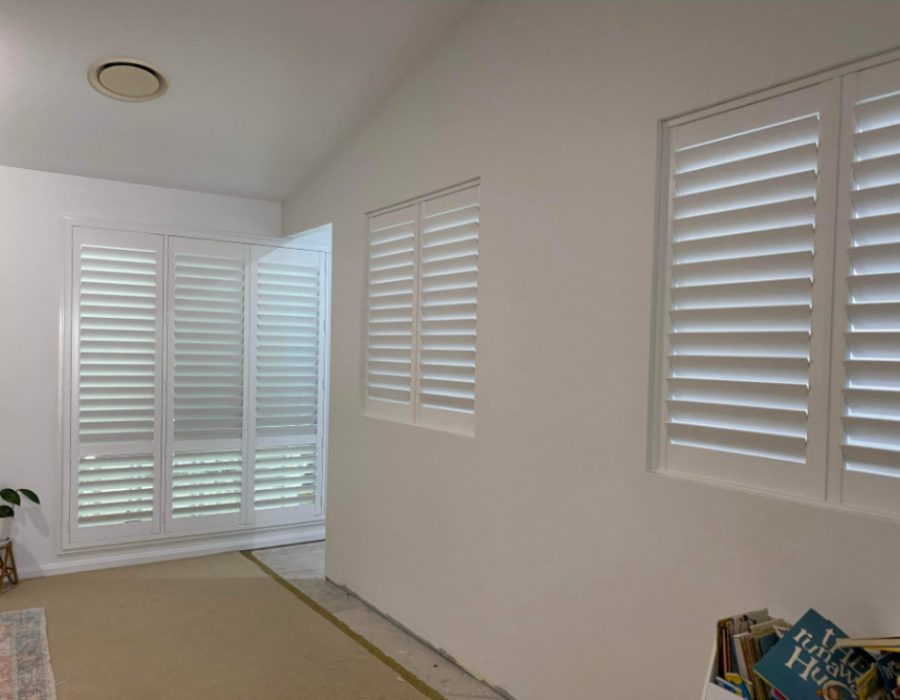 A room with white walls featuring two sets of indoor PVC shutters, allowing light to filter through. A small potted plant sits on the floor in the corner. A stack of books is partially visible on the right.