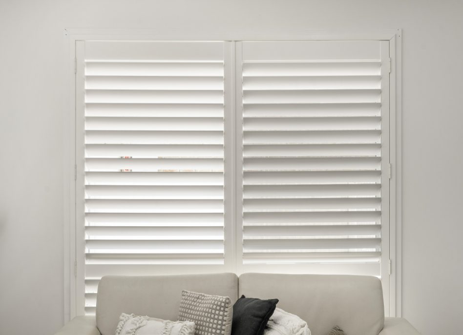 A minimalist living room features a white sofa with several patterned cushions set against large windows adorned with sleek indoor PVC shutters, allowing soft natural light to elegantly filter into the space.